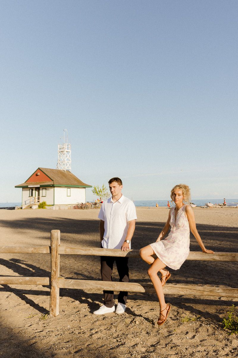 Couple posing near wooden fence on sandy beach with historic lifeguard station at golden hour.