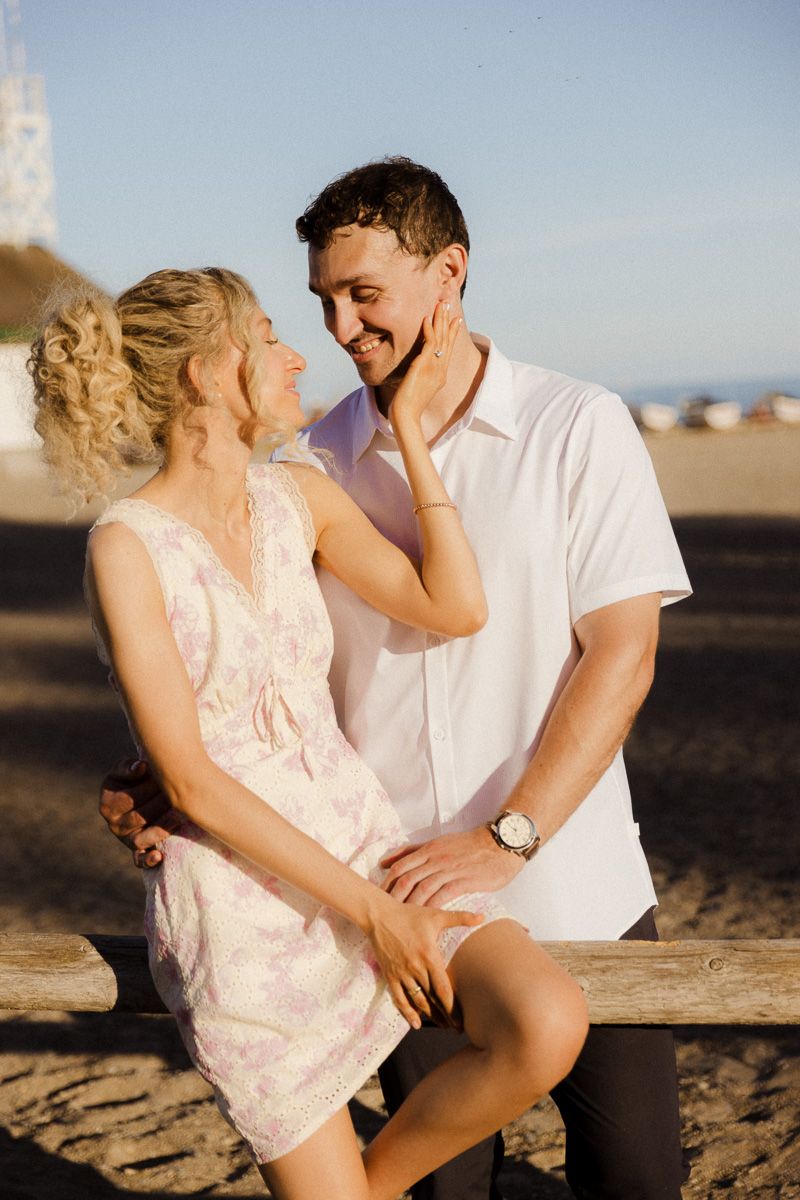 Couple sharing a romantic moment on a beach bench at sunset, woman in floral dress touching man's face.