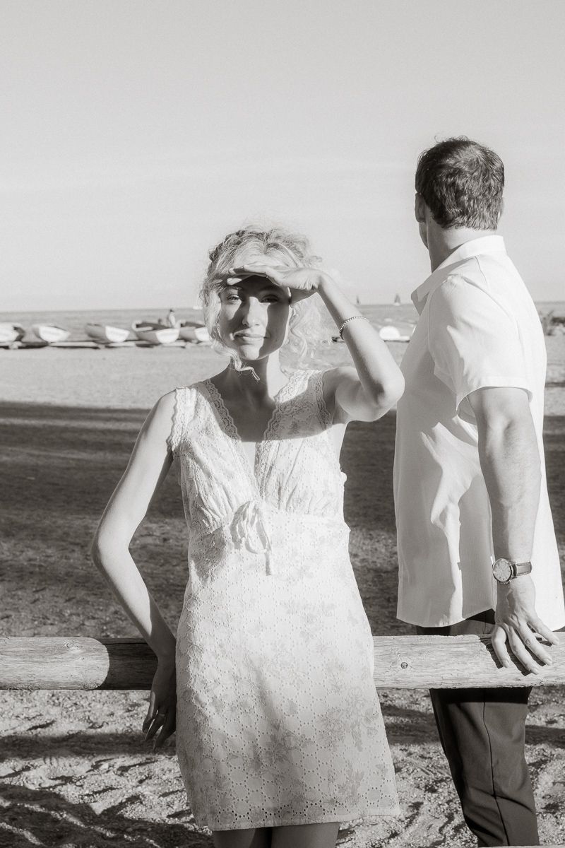 Black and white photo of a blonde woman in white lace dress shielding her eyes on a sunny beach.
