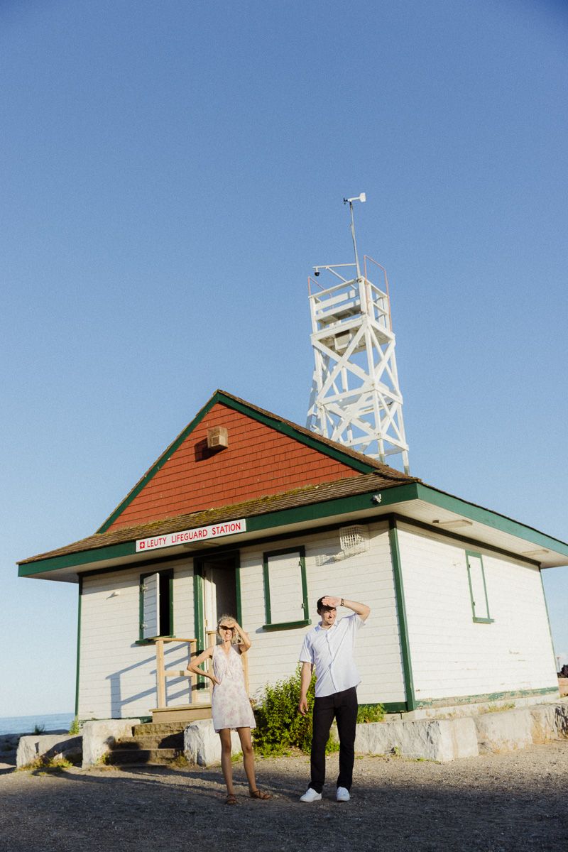Couple standing outside a white and green lifeguard station with a lookout tower under a clear blue sky.