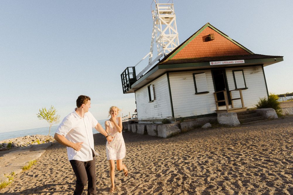 Couple walking hand in hand on sandy beach near a white lighthouse building at golden hour.
