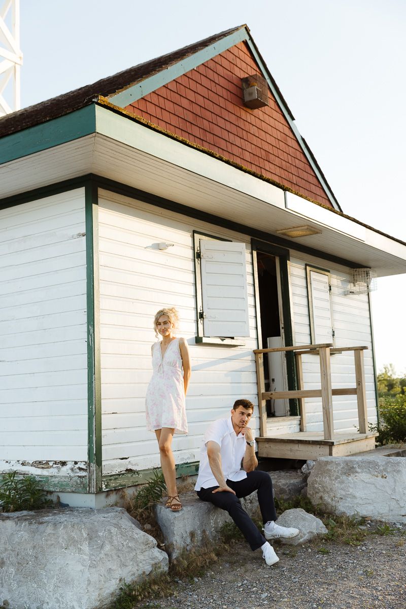 A couple poses outside a white wooden cottage, woman in white dress standing on rocks, man sitting casually nearby.