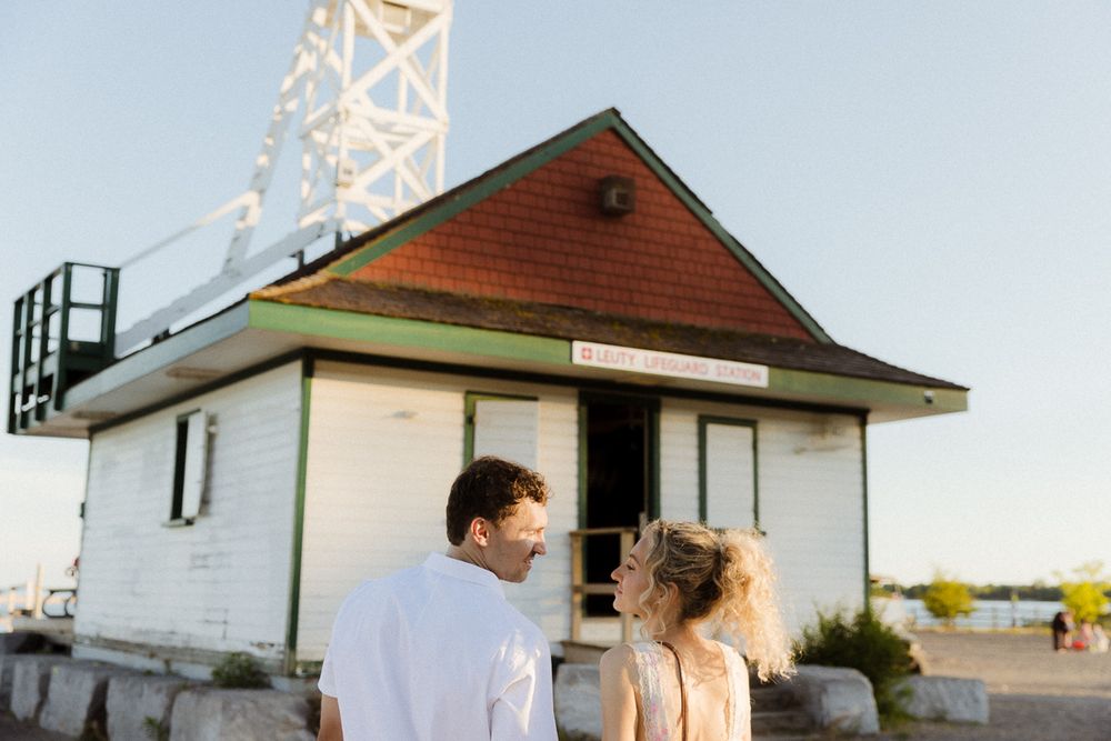 Couple smiling at each other in front of a white lighthouse building at golden hour.