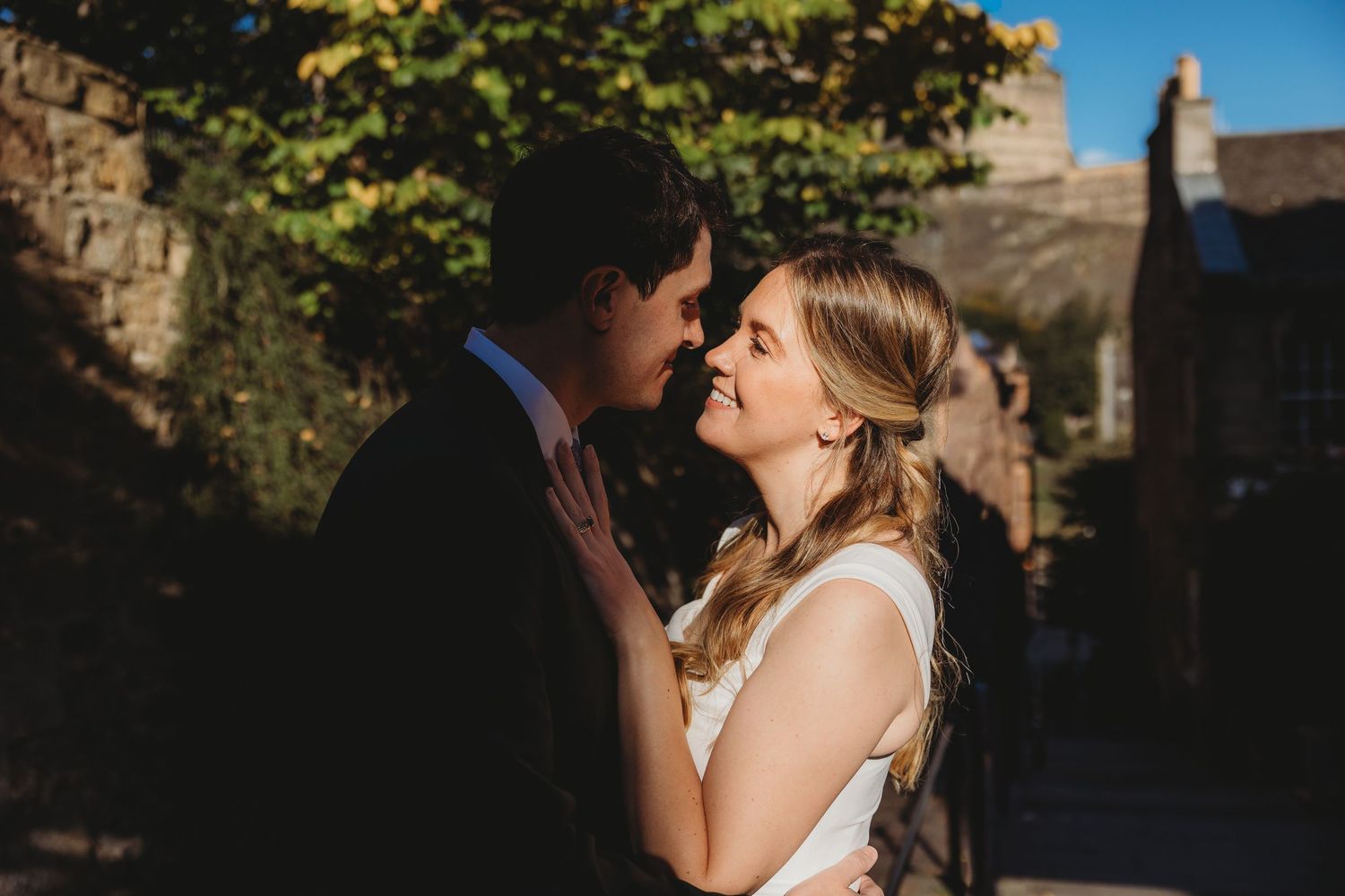 A bride and groom share a romantic moment outdoors, smiling at each other in warm golden sunlight near a stone building.