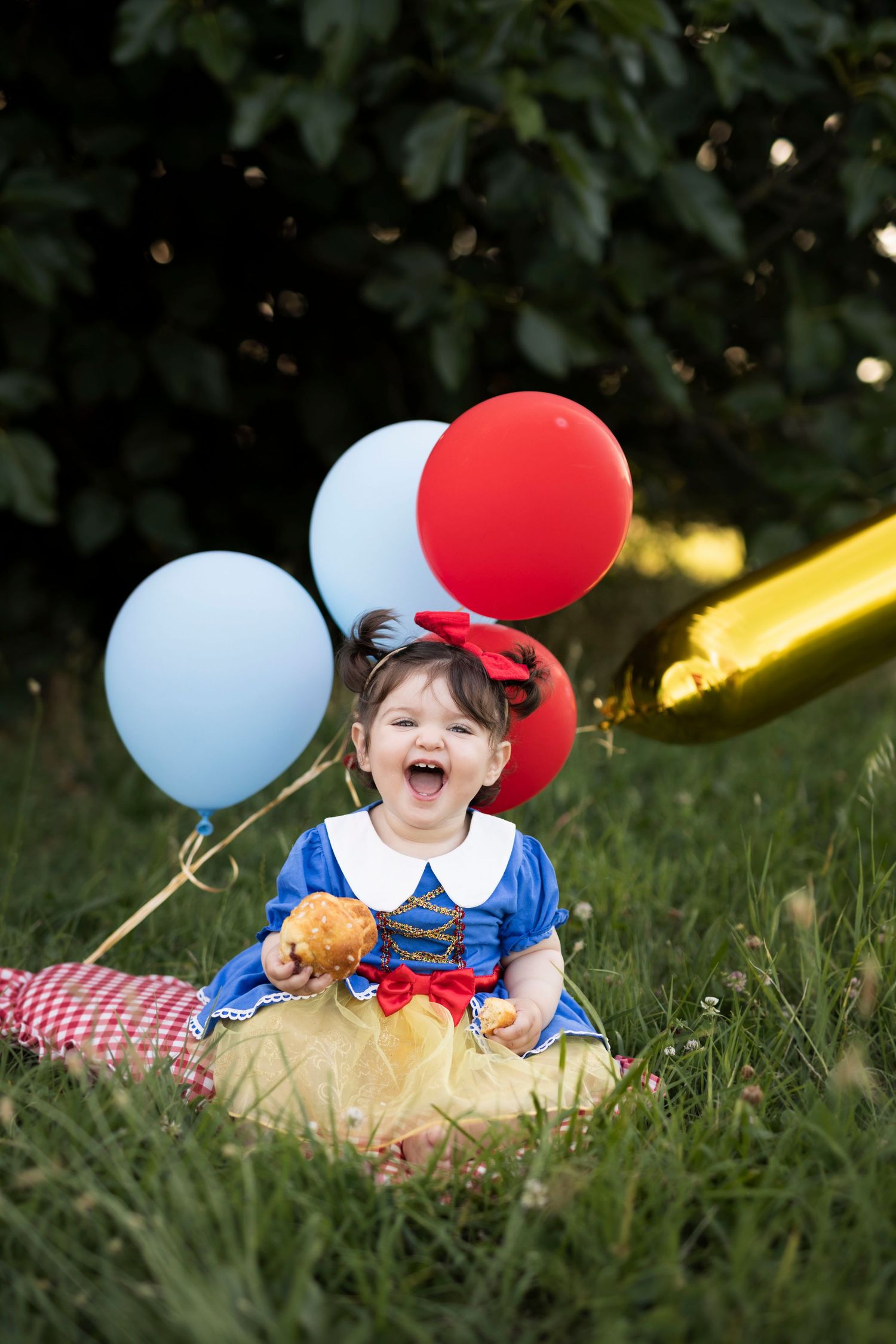 Smiling baby girl in Snow White costume with red bow, yellow tutu, sitting on checkered blanket with red apple outdoors.