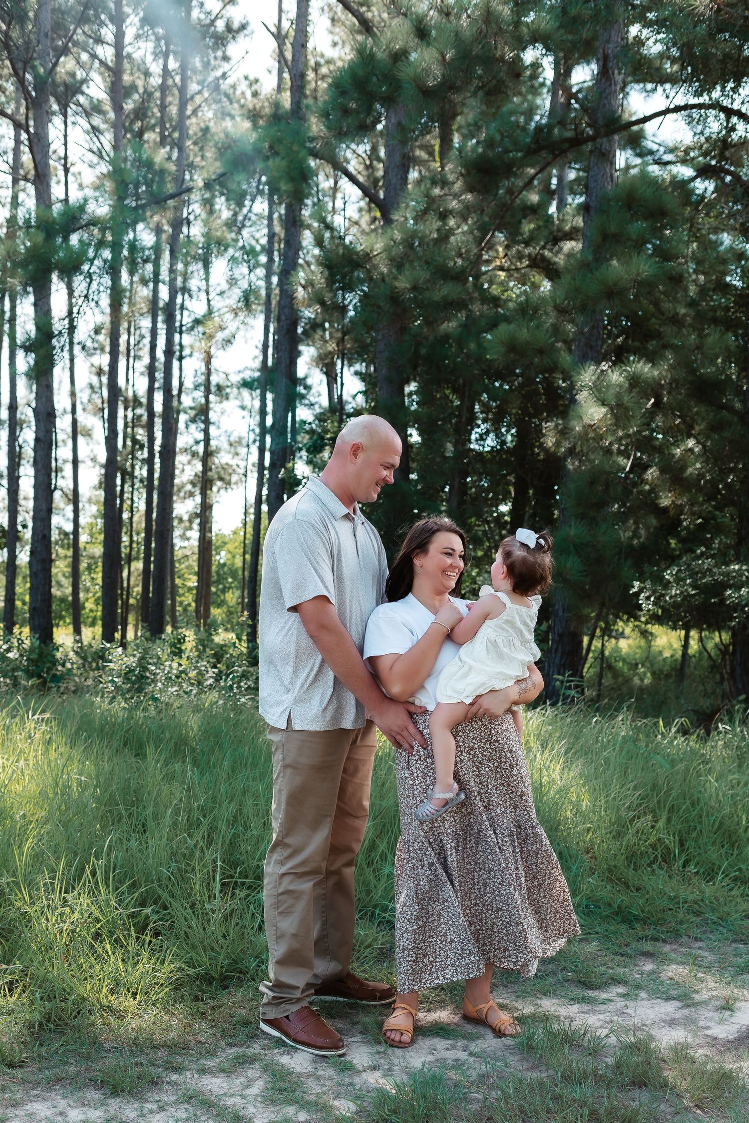 A family of three poses outdoors in a lush pine forest, parents smiling at their young daughter in a sunlit clearing.