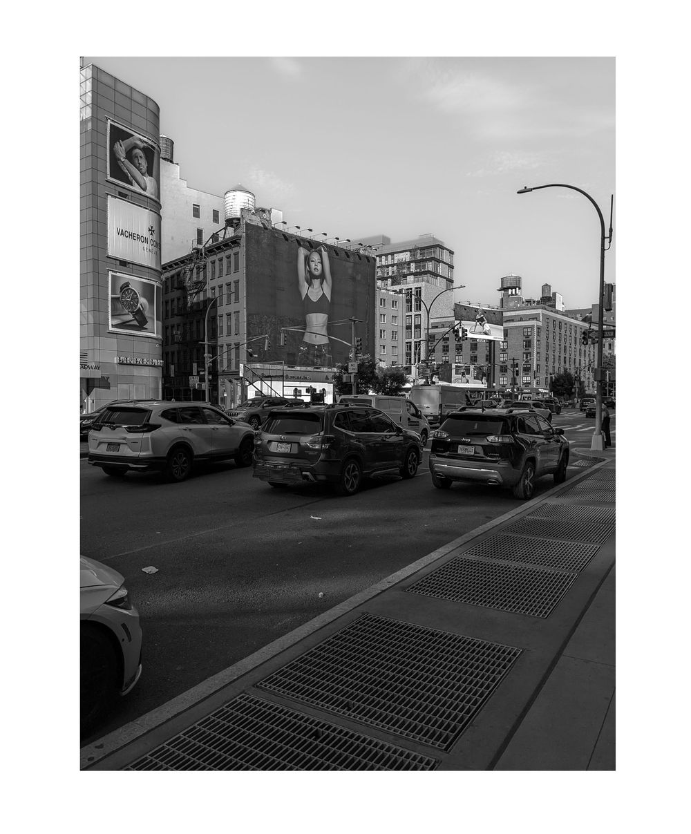 Black and white photo of busy New York City intersection with traffic, large billboards, and urban buildings.