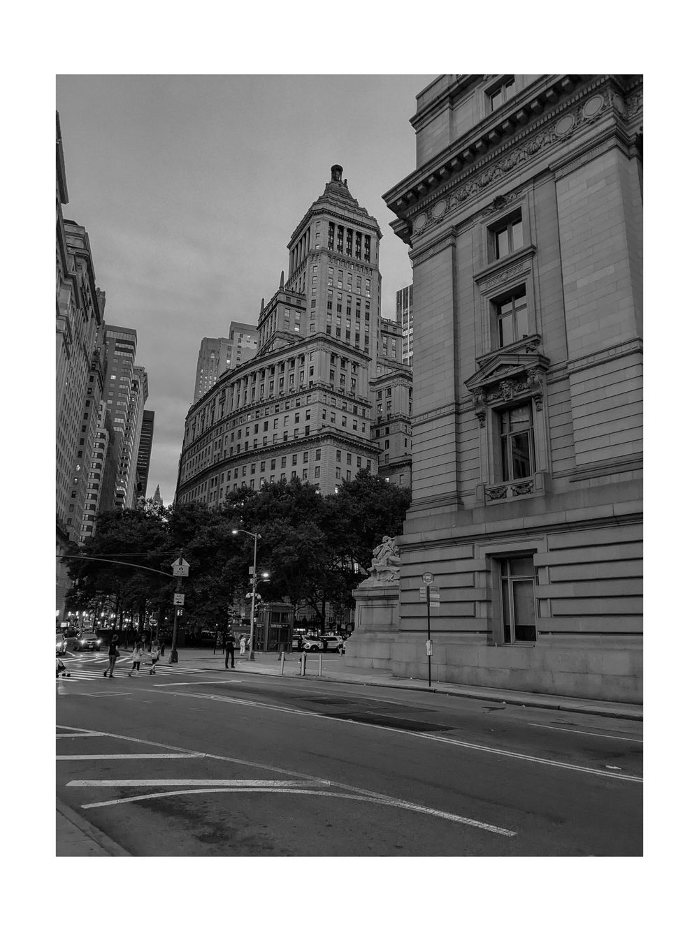 Black and white photo of classic New York City architecture with ornate buildings and tree-lined street under overcast sky.