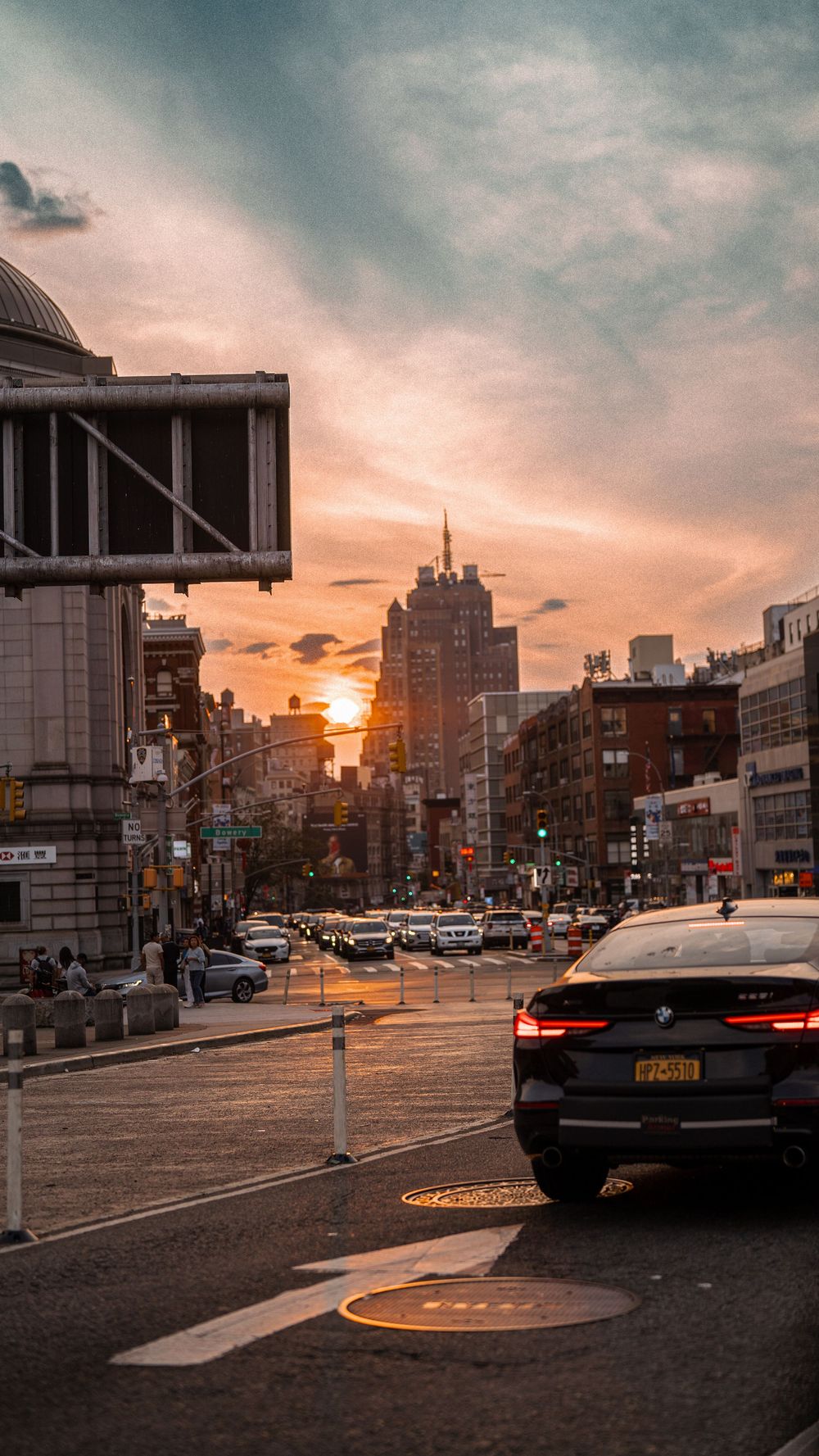 NYC street scene at sunset with BMW car, busy intersection, city buildings, and dramatic orange cloudy sky.