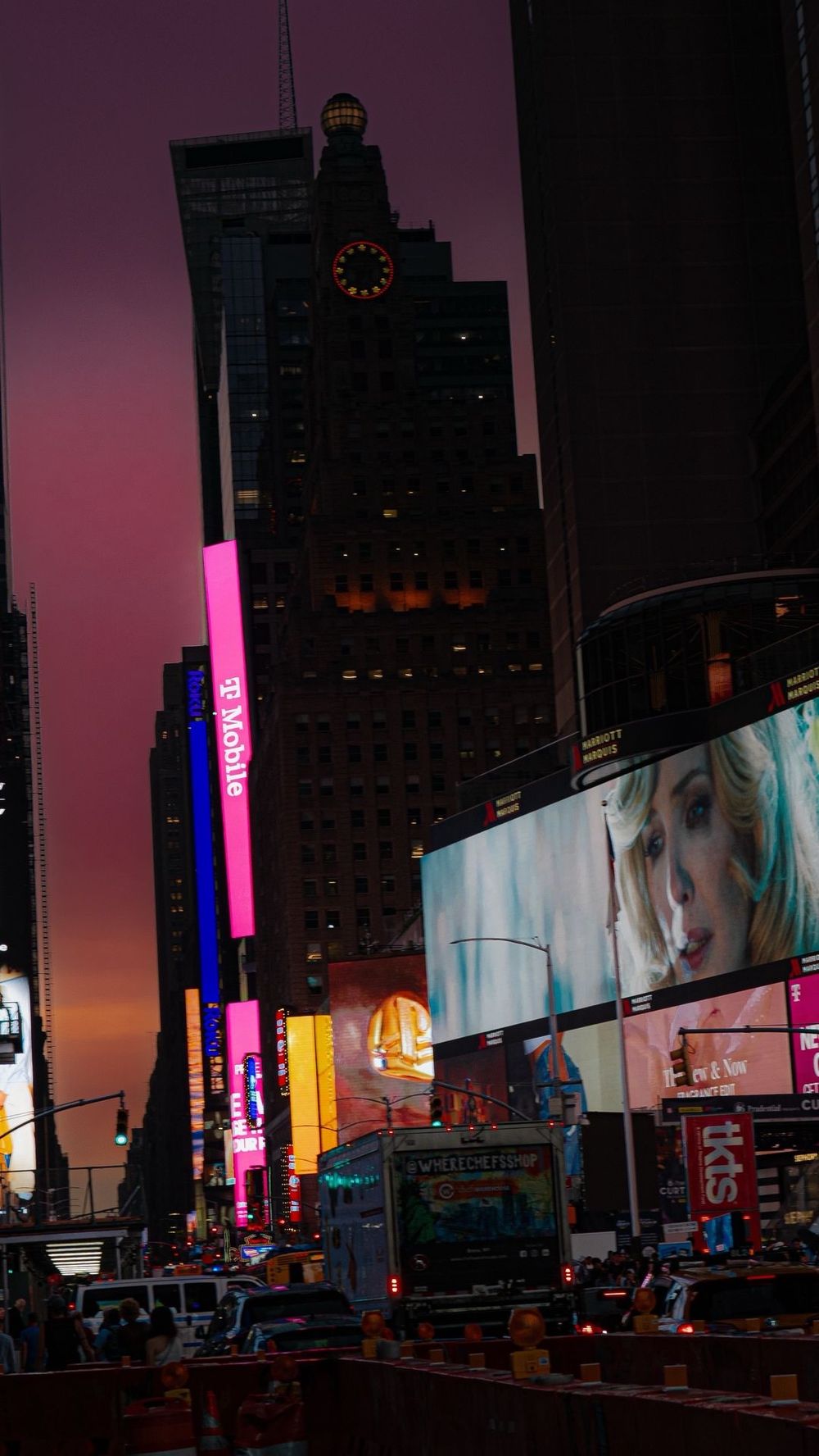 Times Square at night with bright LED billboards, T-Mobile signs, and heavy traffic under a purple sky.