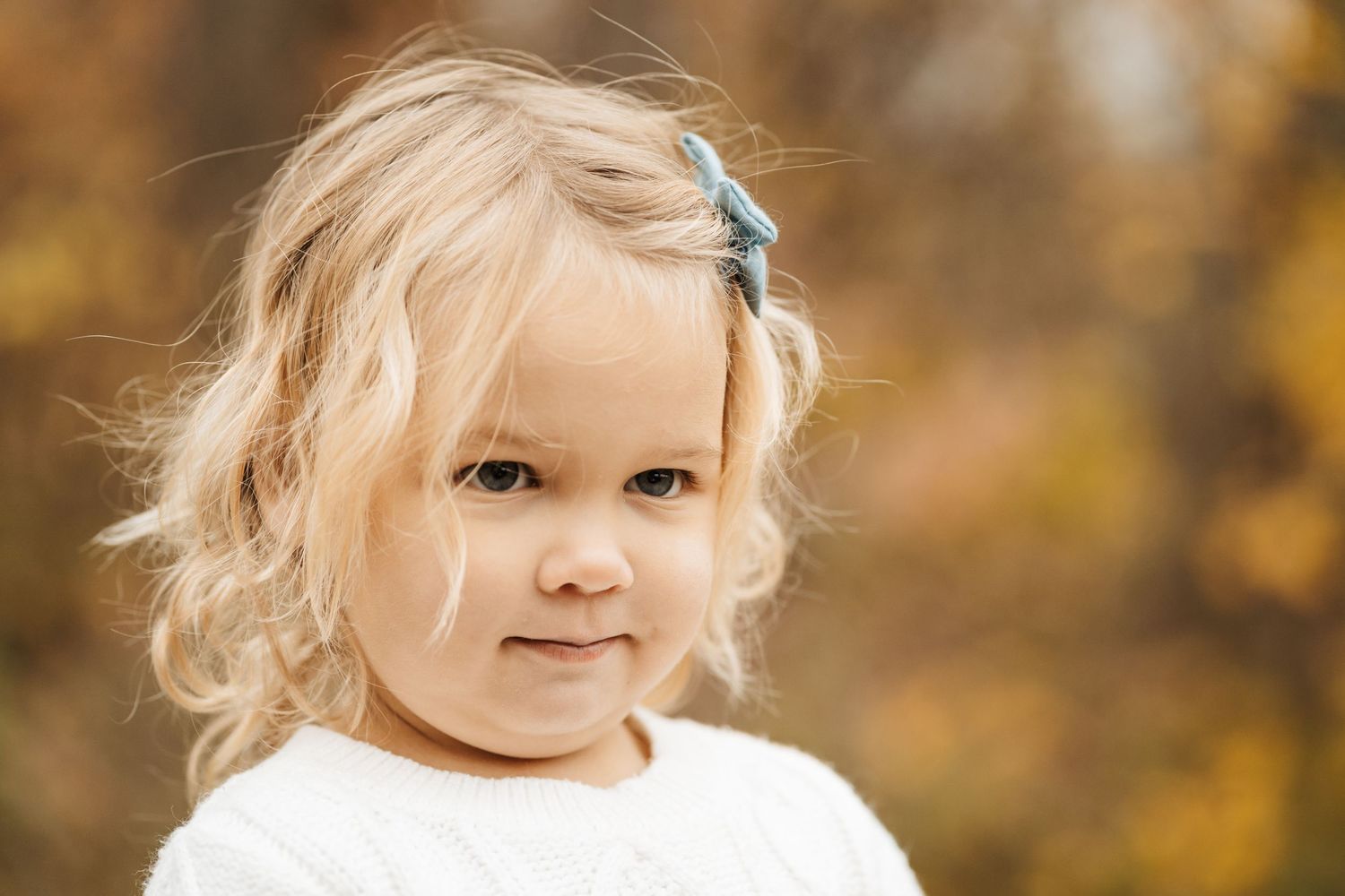 Cute blonde toddler girl with blue hair clip smiling outdoors in autumn setting wearing white sweater.