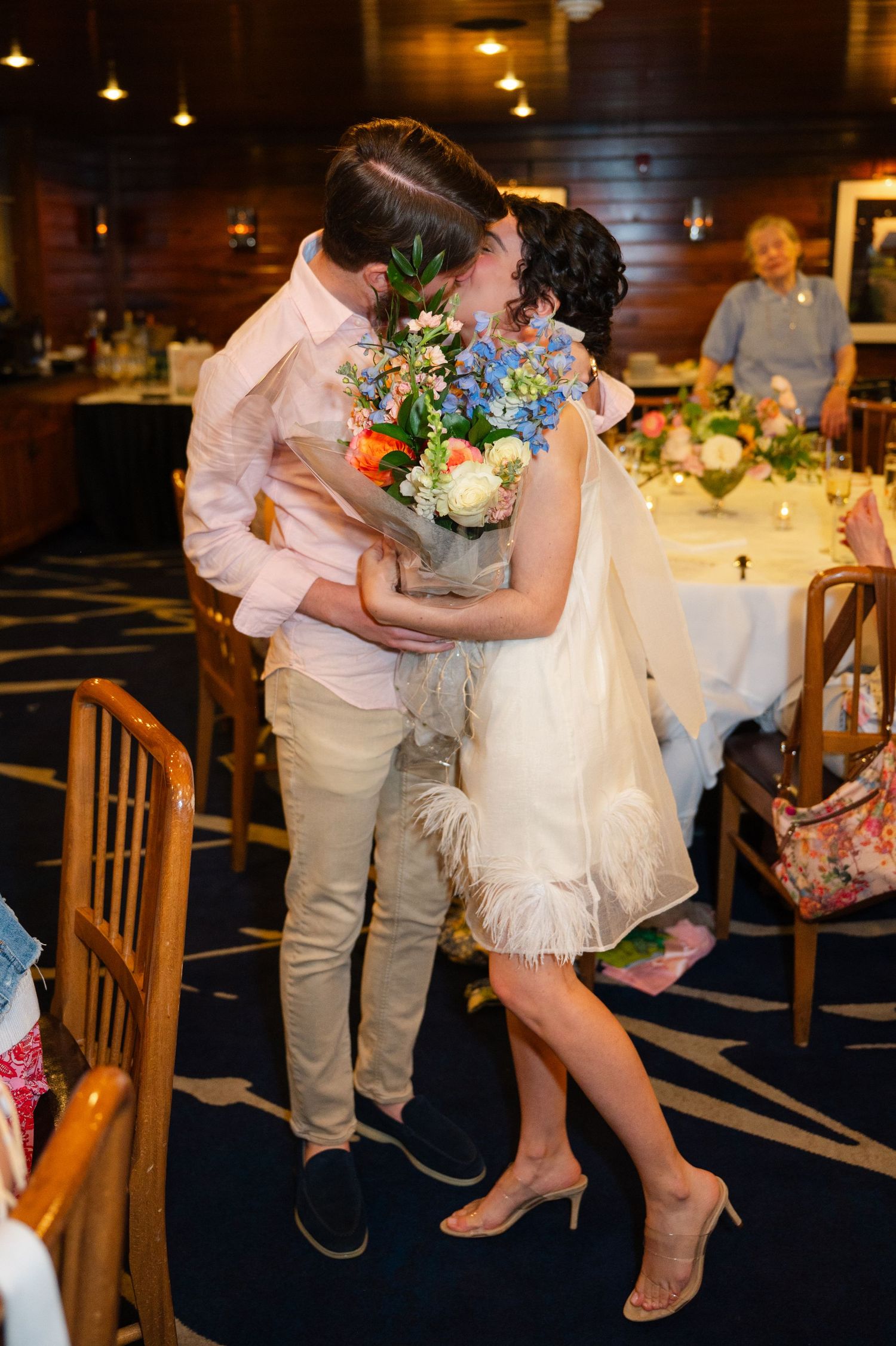 Couple kissing on staircase, woman in white feathered dress holding bouquet of pink and blue flowers.