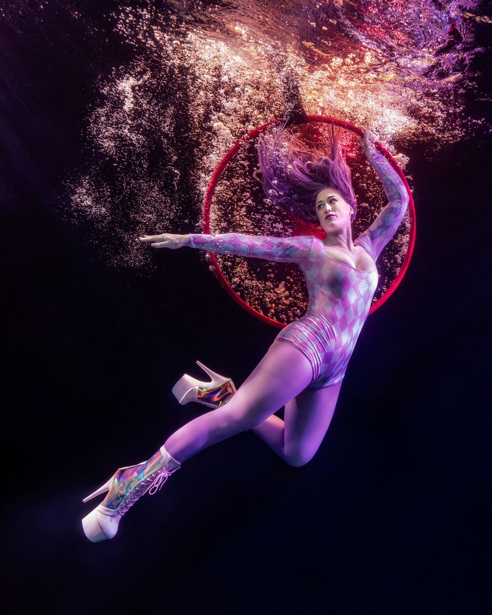 Underwater photo of a woman in purple bodysuit and heels posing gracefully inside a glowing red hoop.