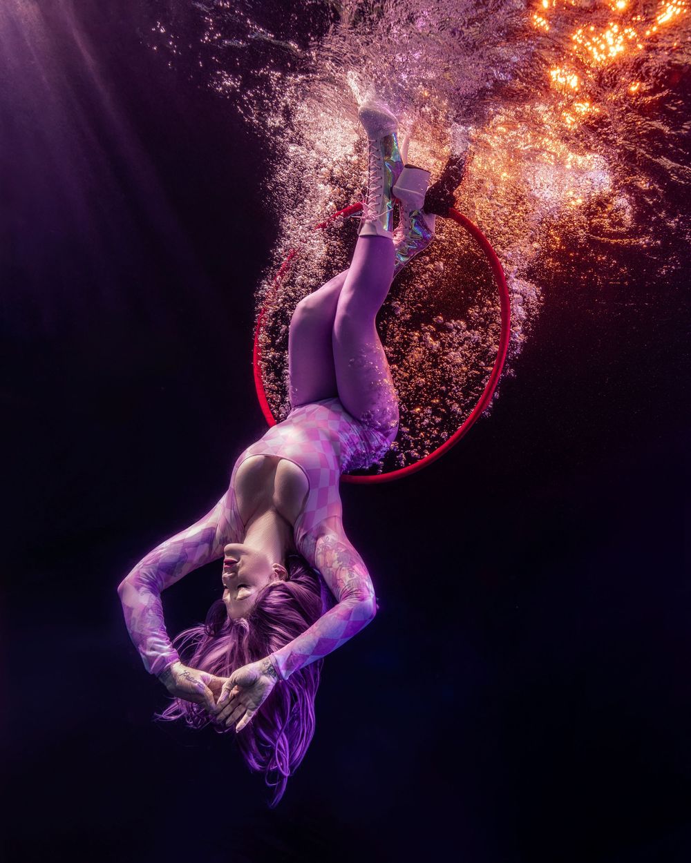 Underwater performer gracefully descending through bubbles with a red hoop, illuminated by dramatic purple and orange light.
