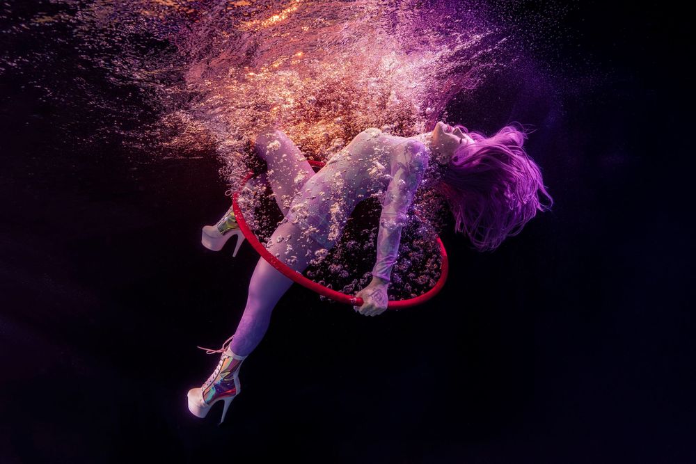 Woman in white dress and heels floating underwater with red hoop, surrounded by purple bubbles on dark background.
