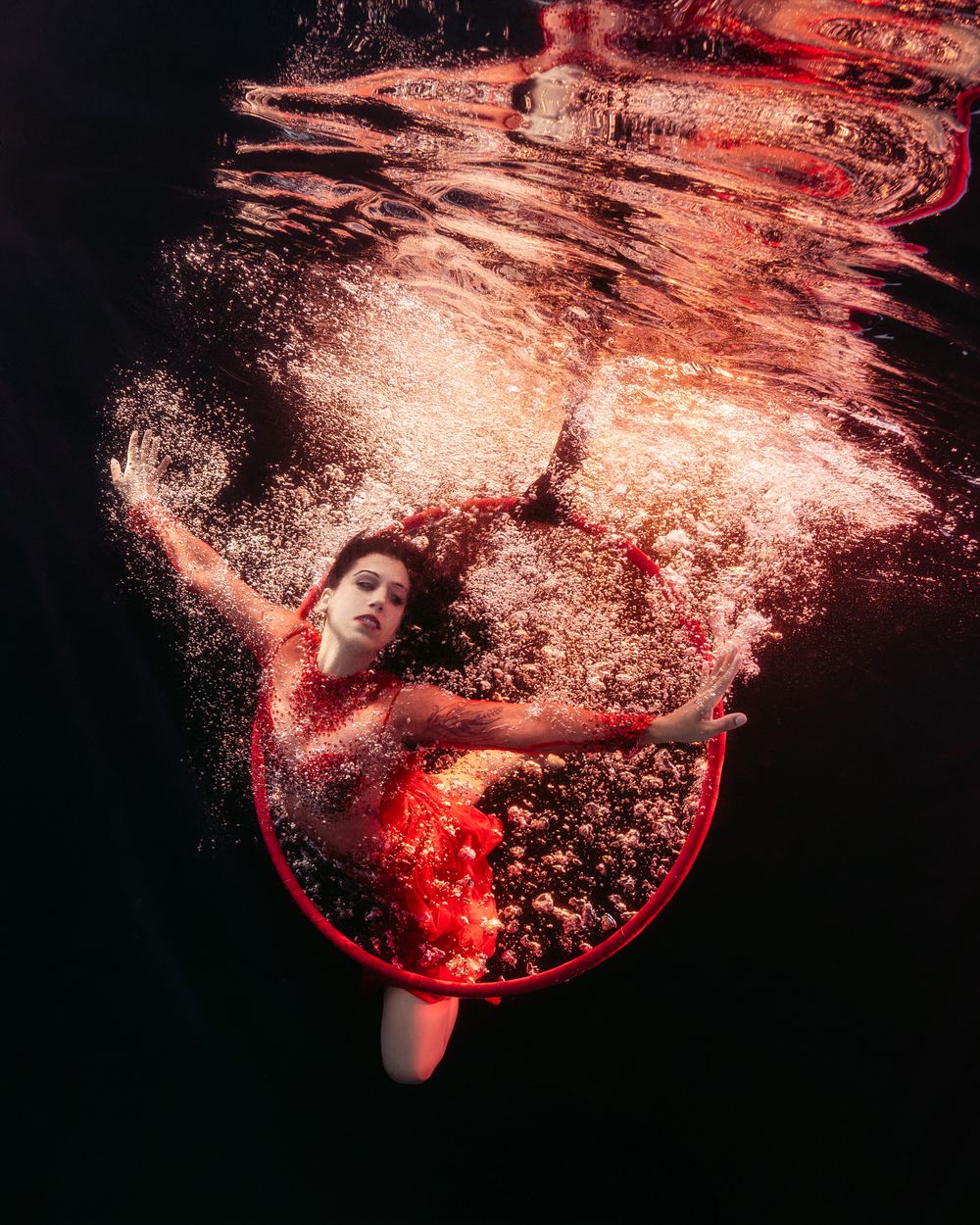 Woman in red dress gracefully swimming underwater with a red hoop, surrounded by bubbles and dramatic lighting.
