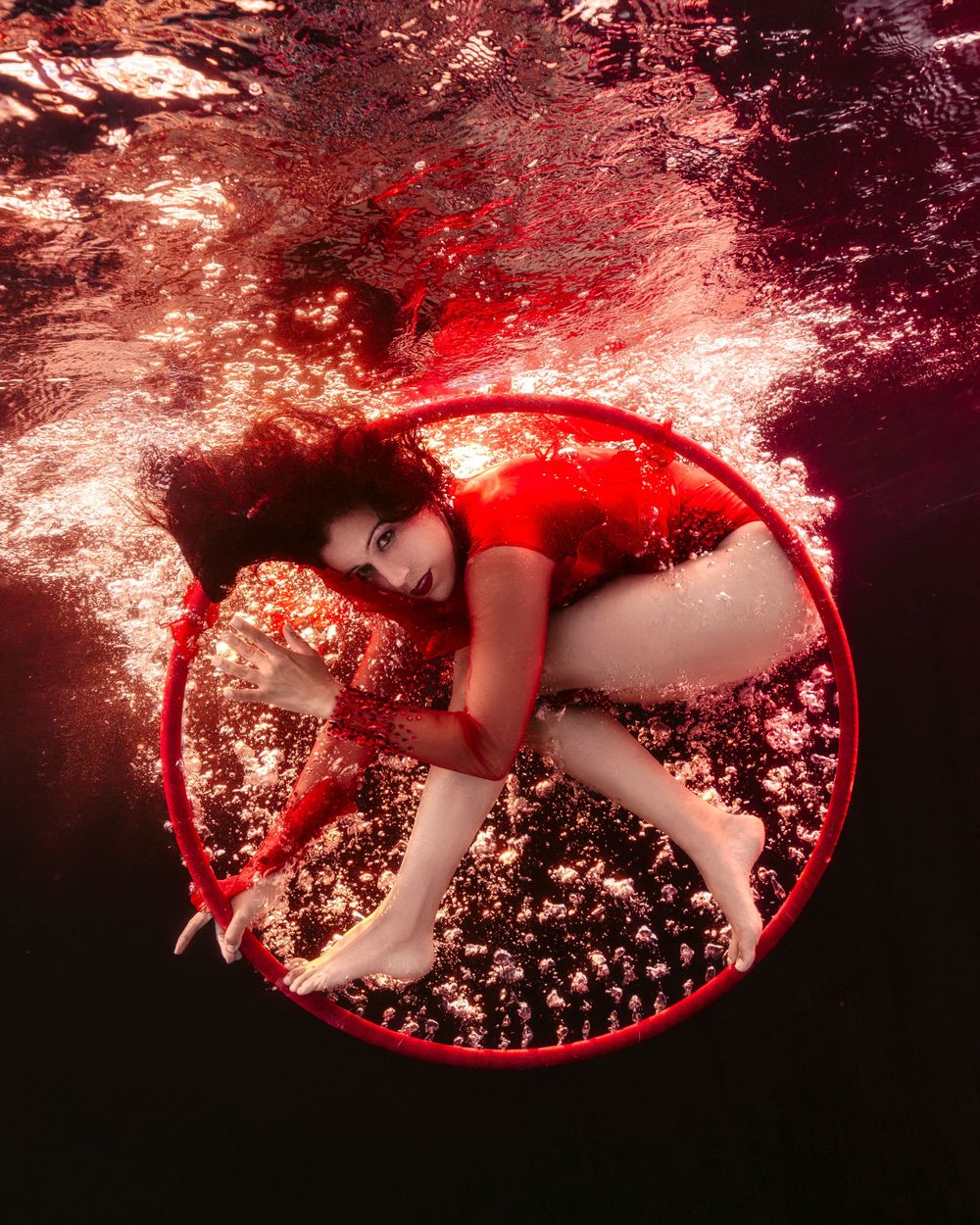Woman in red dress poses gracefully inside a red hoop underwater, surrounded by bubbles and dramatic red lighting.