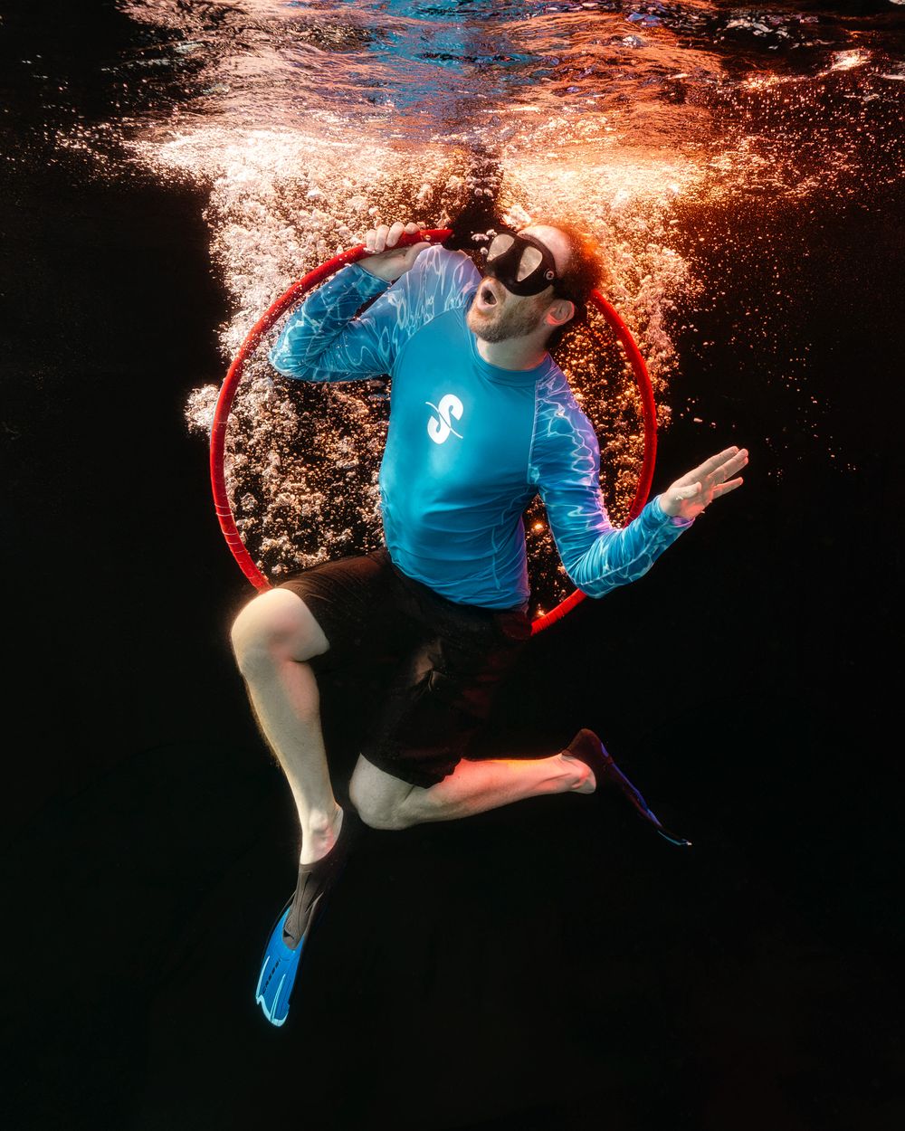 Underwater freediver in blue rash guard, black shorts, and blue fins poses inside a red hoop with bubbles rising above.