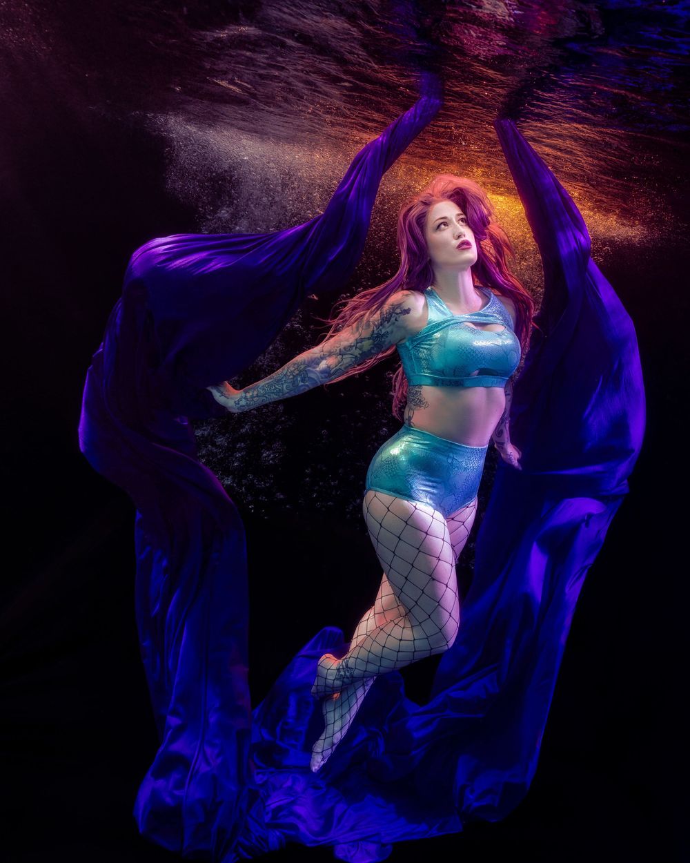 Underwater photo of a woman with red hair in a teal outfit, surrounded by flowing purple fabric against a dark background.