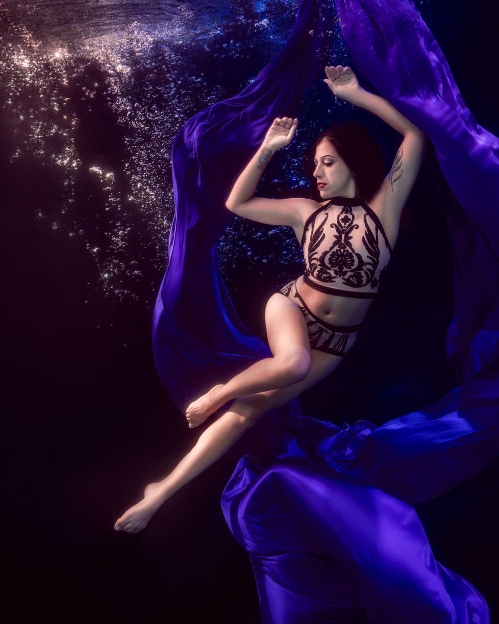 Woman in black lace lingerie floating underwater surrounded by flowing purple fabric and bubbles in dark water.