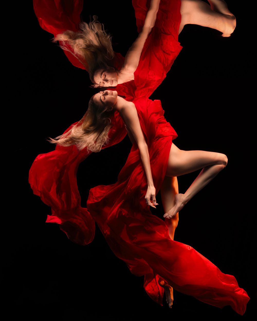 Two women in flowing red gowns dance gracefully against a black background, their fabric swirling elegantly.