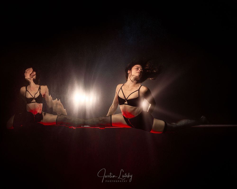 Two women in lingerie pose dramatically on a red surface with bright backlighting in a dark, moody boudoir photography setting.