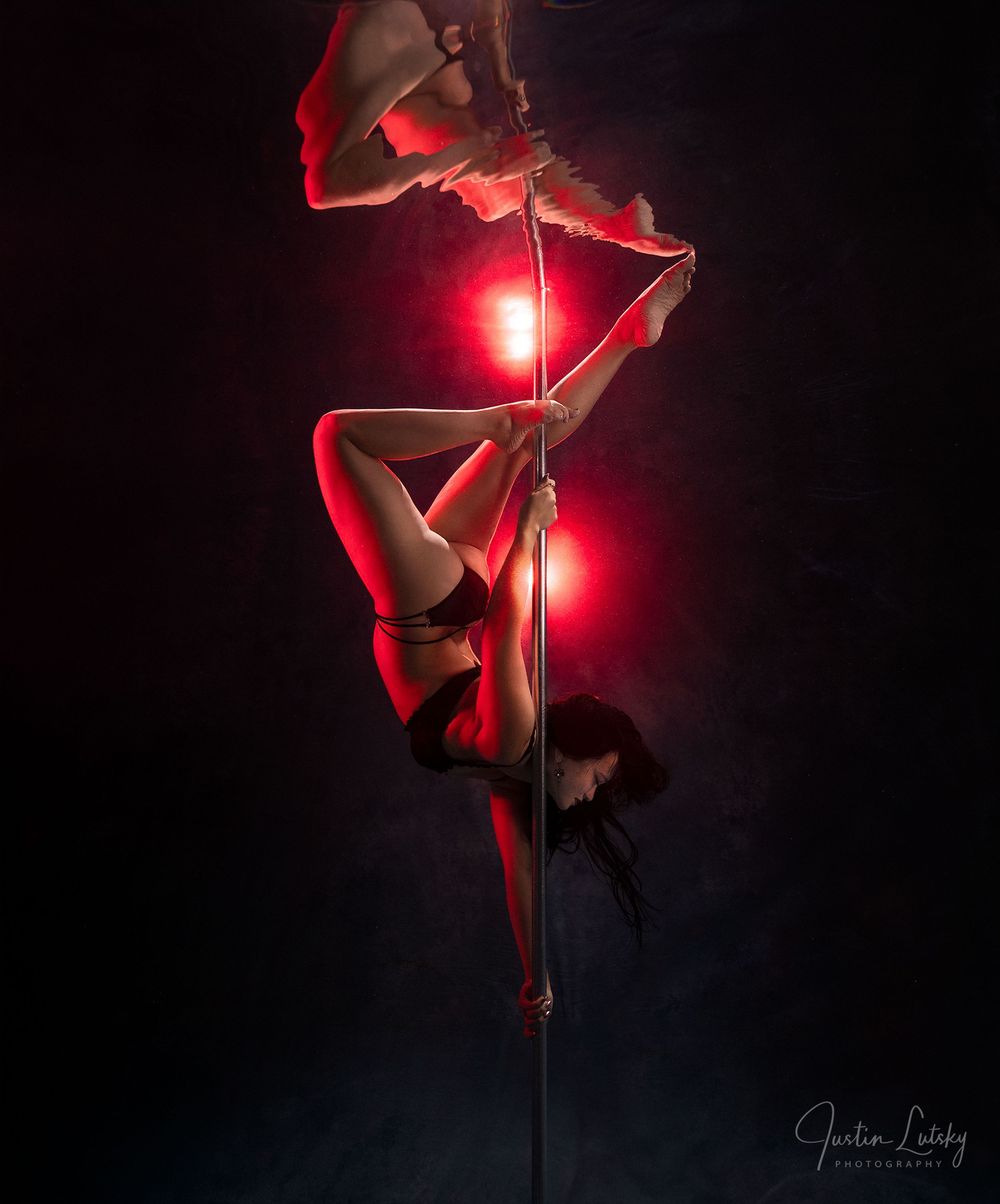 Aerial pole dancer performs an inverted split under dramatic red lighting against a dark background.