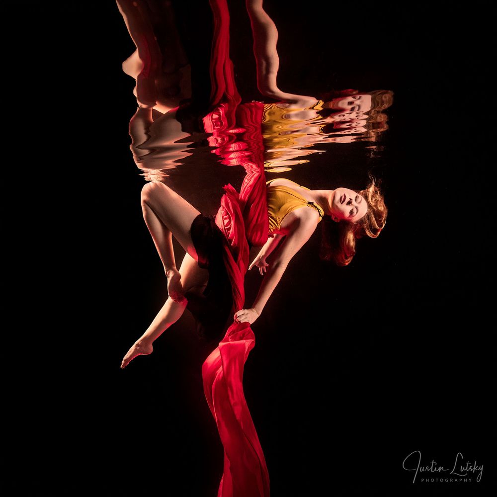 Underwater photo of a dancer in red dress gracefully posed, with a mirror reflection on the water surface against black background.