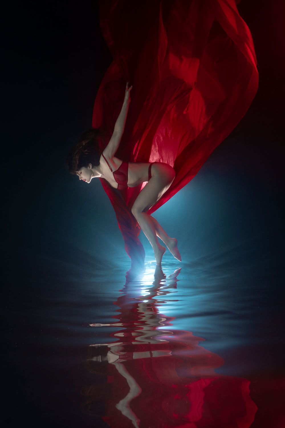 Woman in flowing red fabric stands in dark water with blue light reflection creating dramatic artistic scene.