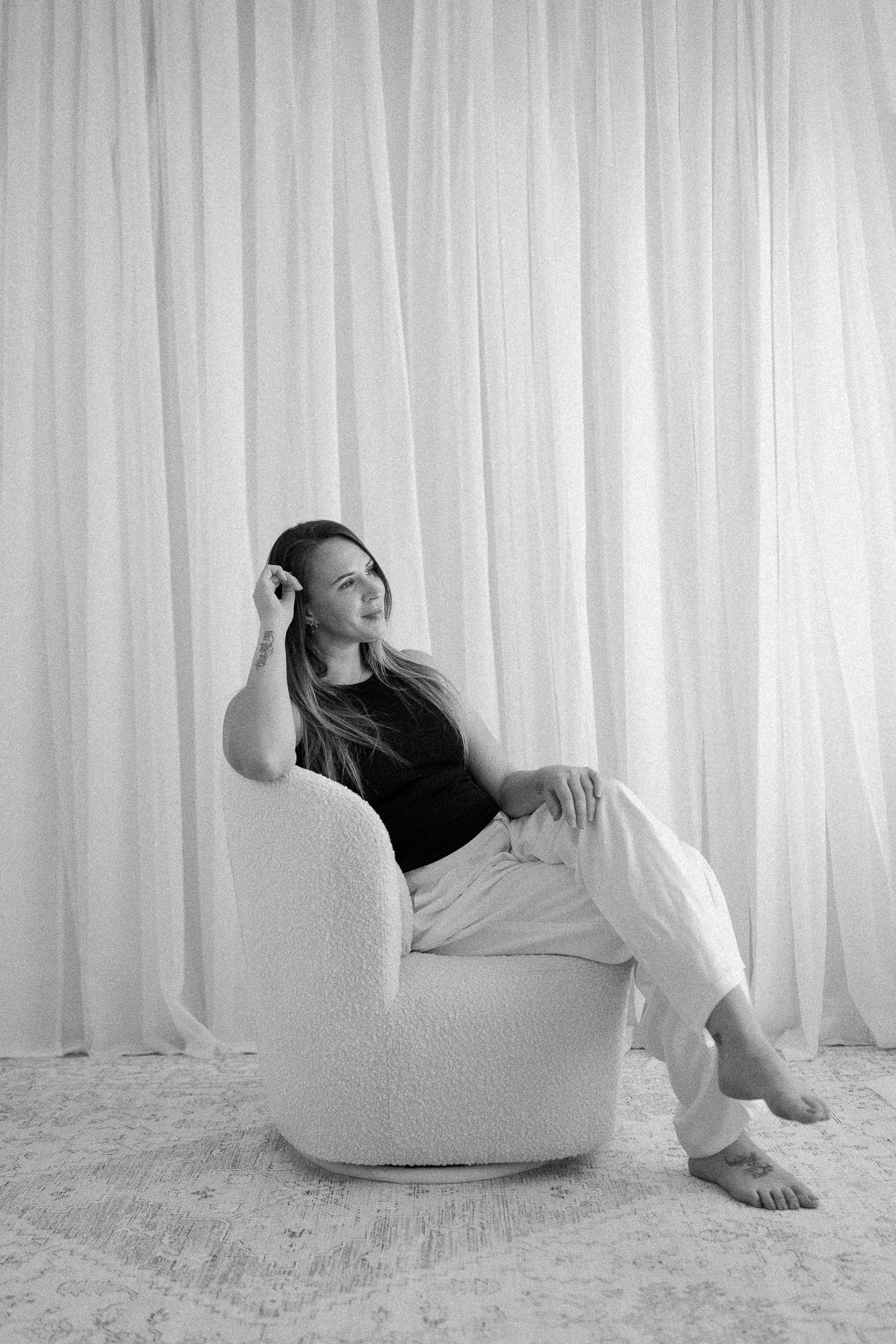 Black and white photo of a woman sitting relaxed in a fluffy white chair against sheer curtains.