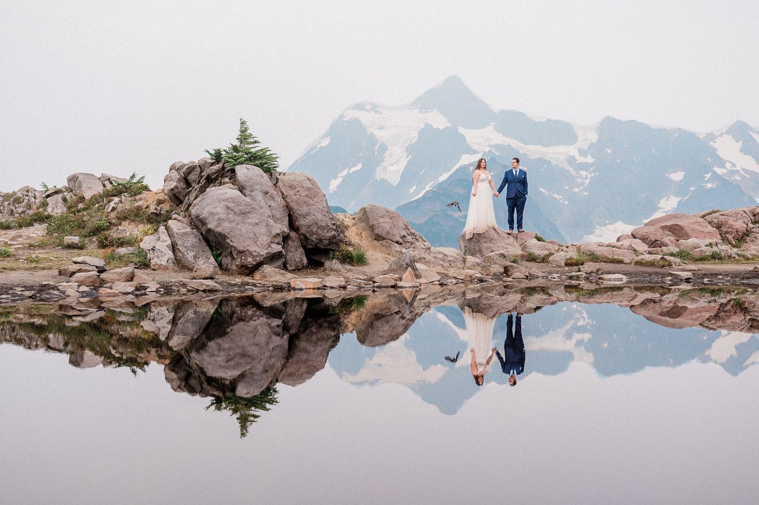 Couple stands on rocky mountain shore with stunning snowy peak reflection in calm alpine lake during outdoor elopement at Artist Point.