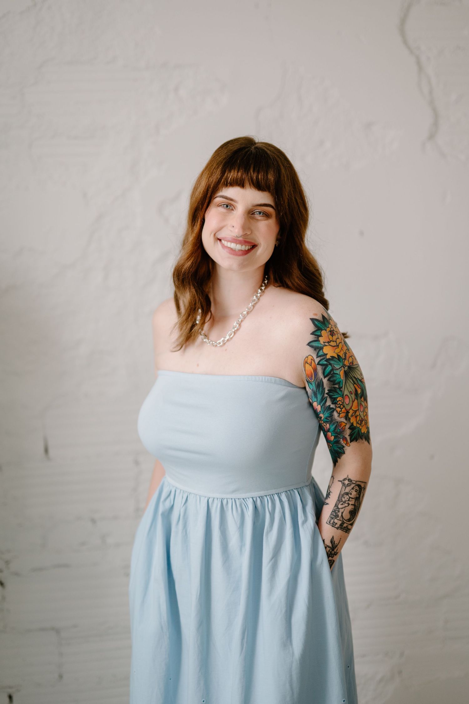 Smiling woman with bangs in light blue strapless dress and floral arm tattoo poses against white brick wall.