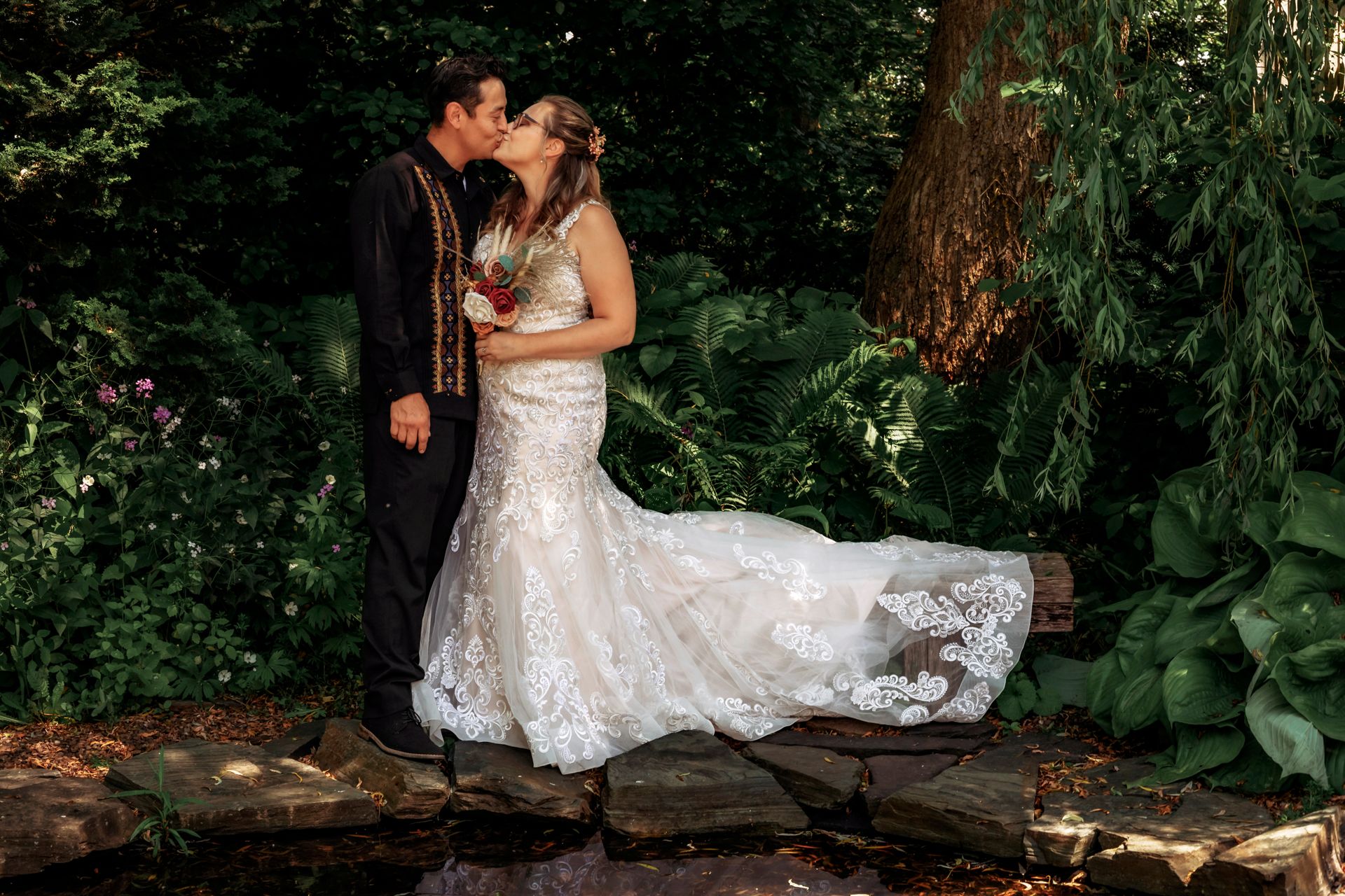 Bride and groom share a romantic kiss in a lush garden setting, bride wearing elegant lace mermaid wedding gown.