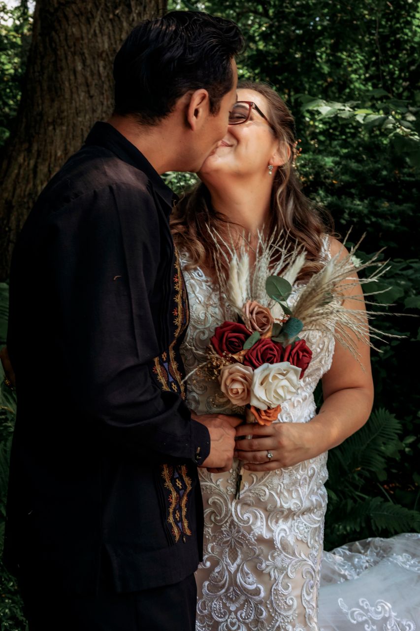 Bride and groom share a romantic kiss outdoors, bride holding a bouquet of red, pink, and white roses with dried pampas grass.