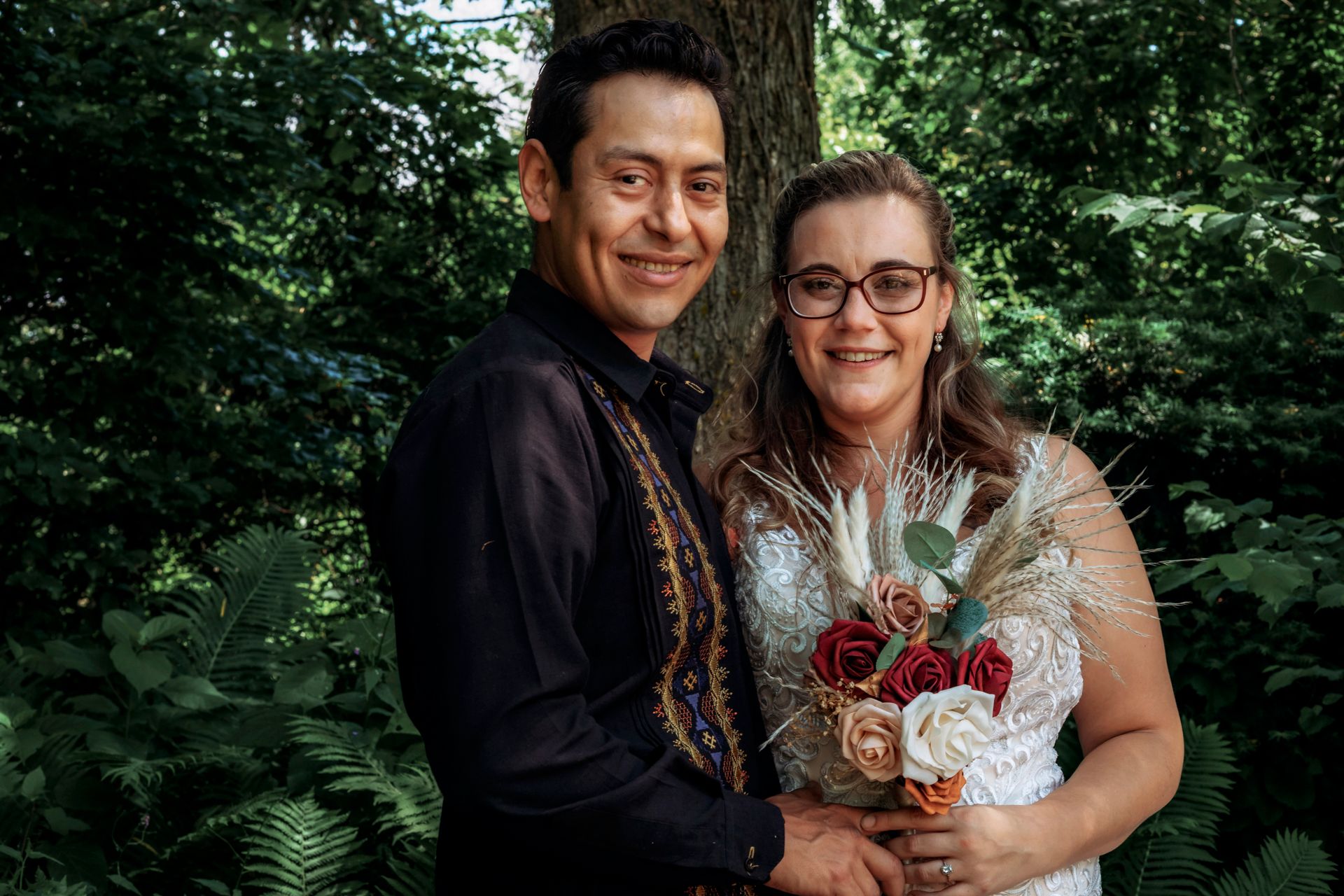 Smiling couple in wedding attire posing outdoors among lush green ferns and trees.