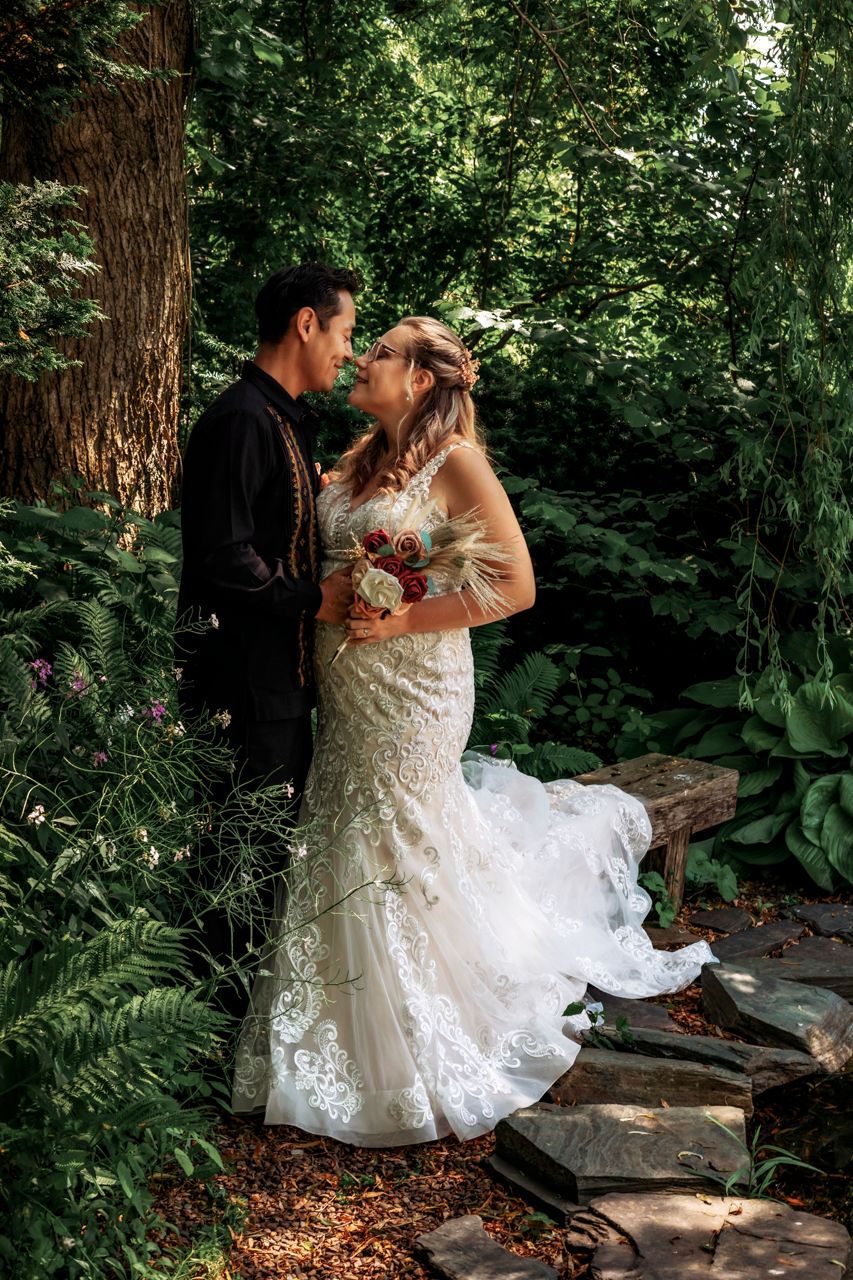 Bride in lace gown and groom in black suit share romantic moment in lush garden setting on their wedding day.