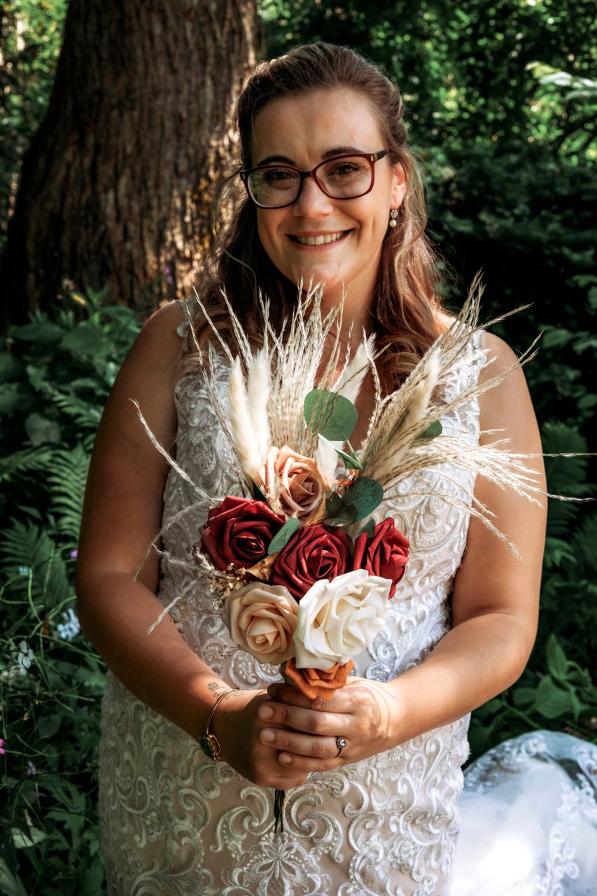 Smiling bride in white lace dress holding bouquet of red, cream roses and pampas grass in lush green forest setting.