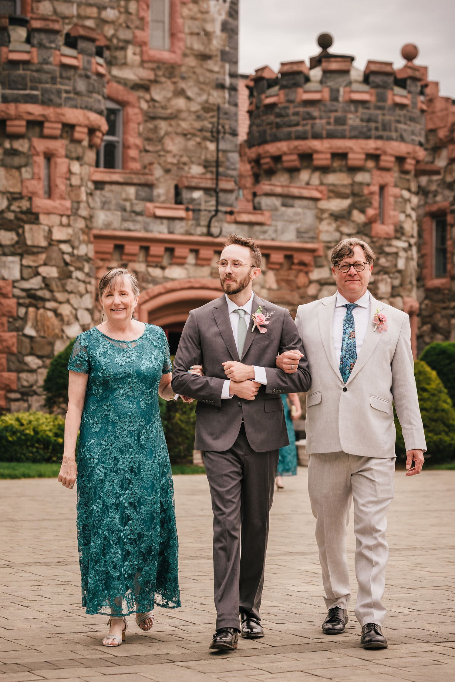Groom walking arm in arm with his parents toward the ceremony at Searles Castle, castle towers behind them.