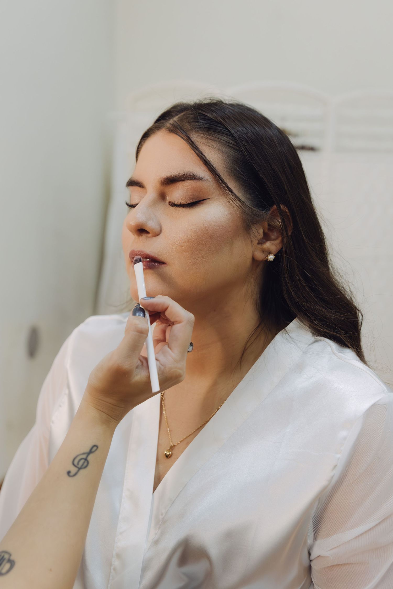 Young woman in white robe applying lip gloss with eyes closed during bridal makeup preparation.