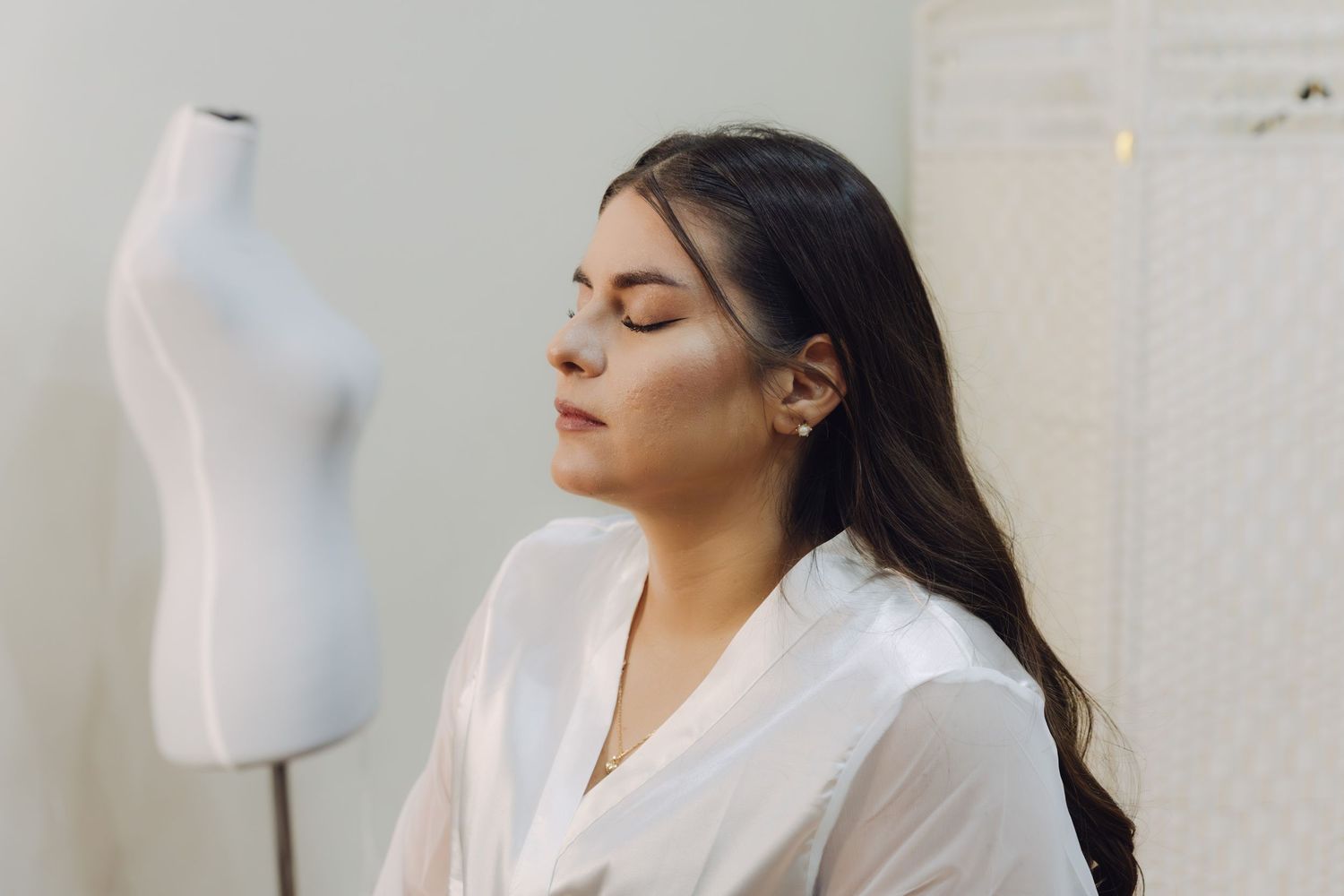 Serene young woman in white silk robe with eyes closed sitting near white dress form mannequin.