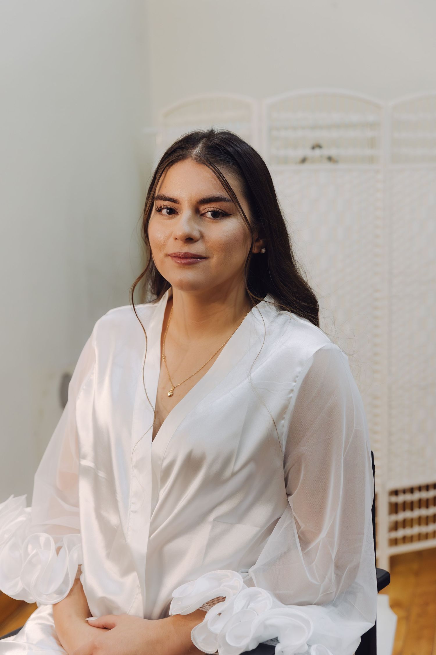 Beautiful young bride in white satin robe sitting calmly smiling during wedding day preparations.