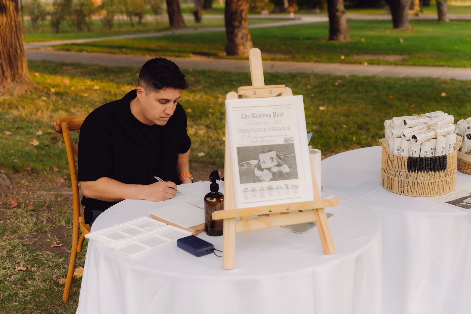 Man in black shirt writing calligraphy at white table with easel sign at outdoor wedding reception.