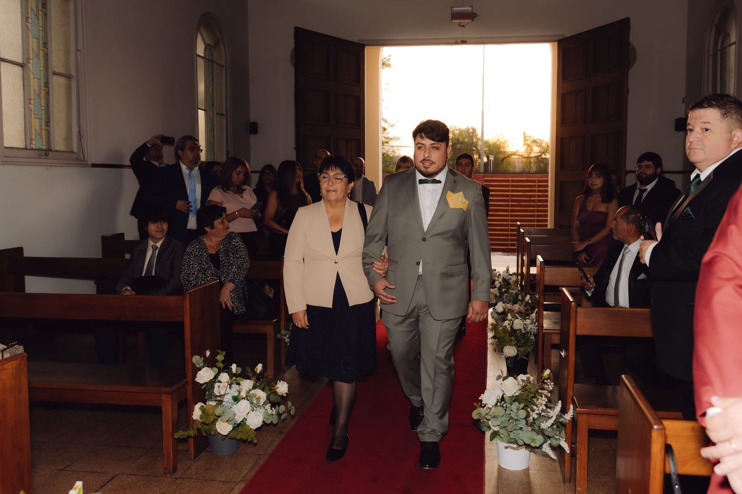 Groom in gray suit with bow tie walks down red carpet aisle with mother at church wedding.