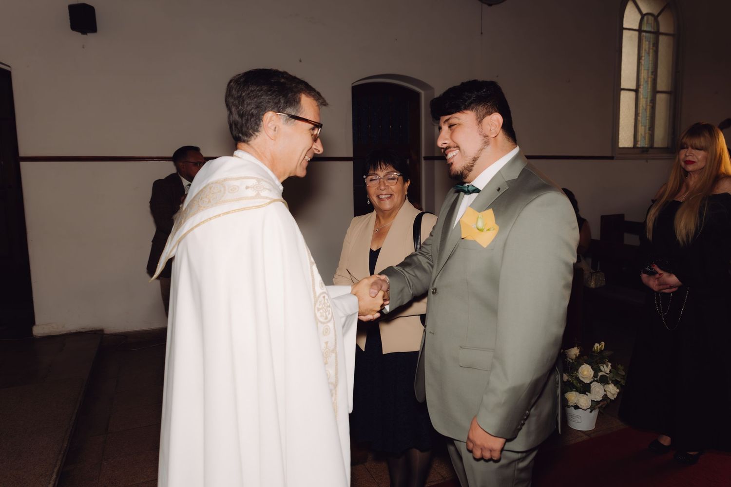 Groom in gray suit shakes hands with Catholic priest in white vestments at church altar.