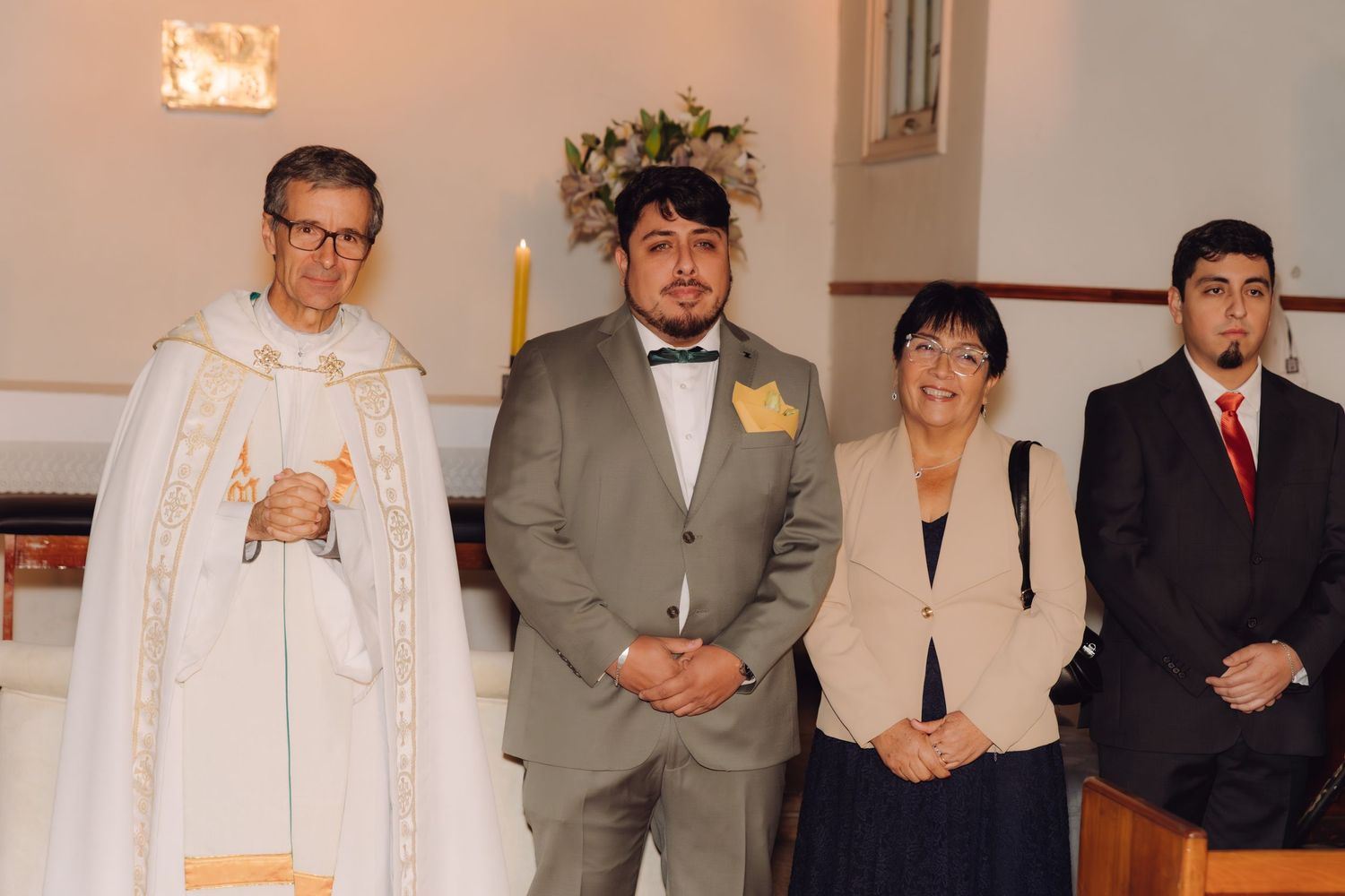 Groom in gray suit poses with priest in white vestments and family members at church altar.