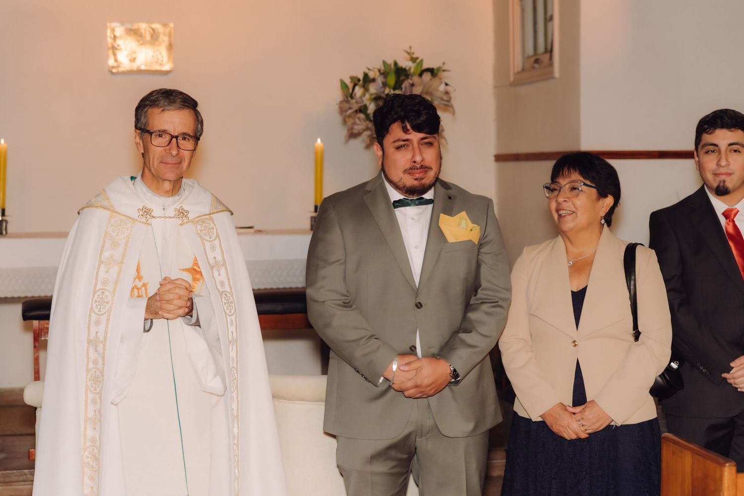 Groom in gray bow tie suit stands with priest and mother at church wedding ceremony.