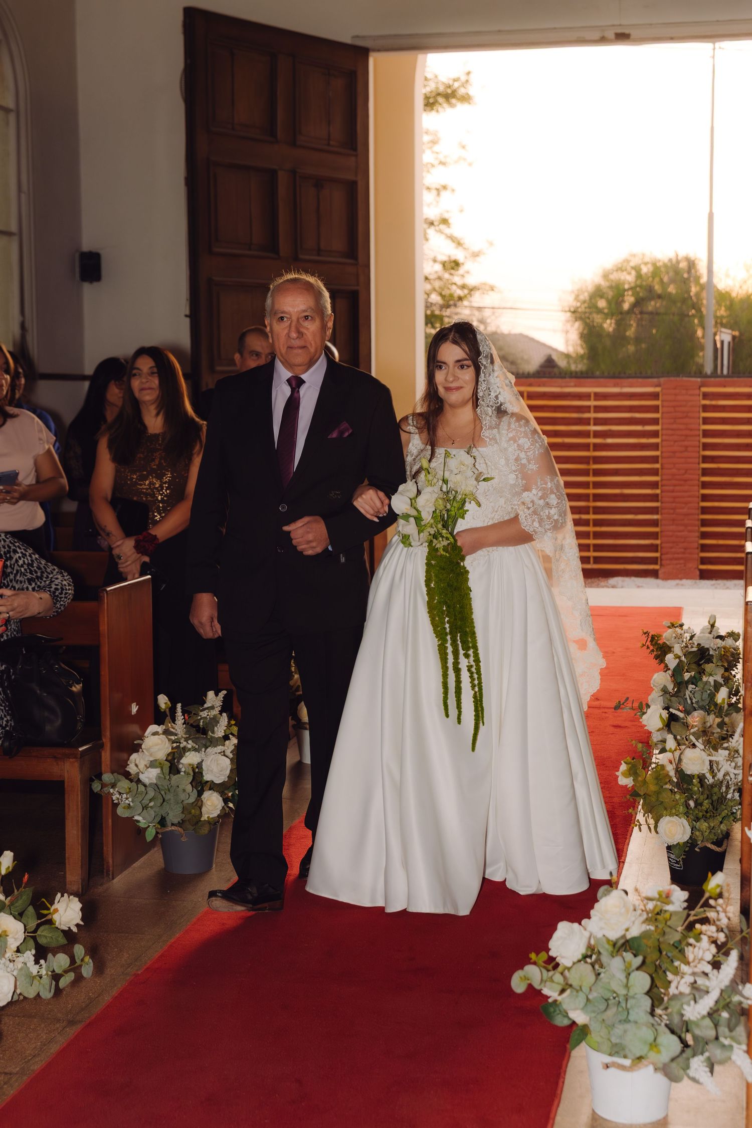 Bride in white lace gown with veil walks down red carpet aisle escorted by elderly man.
