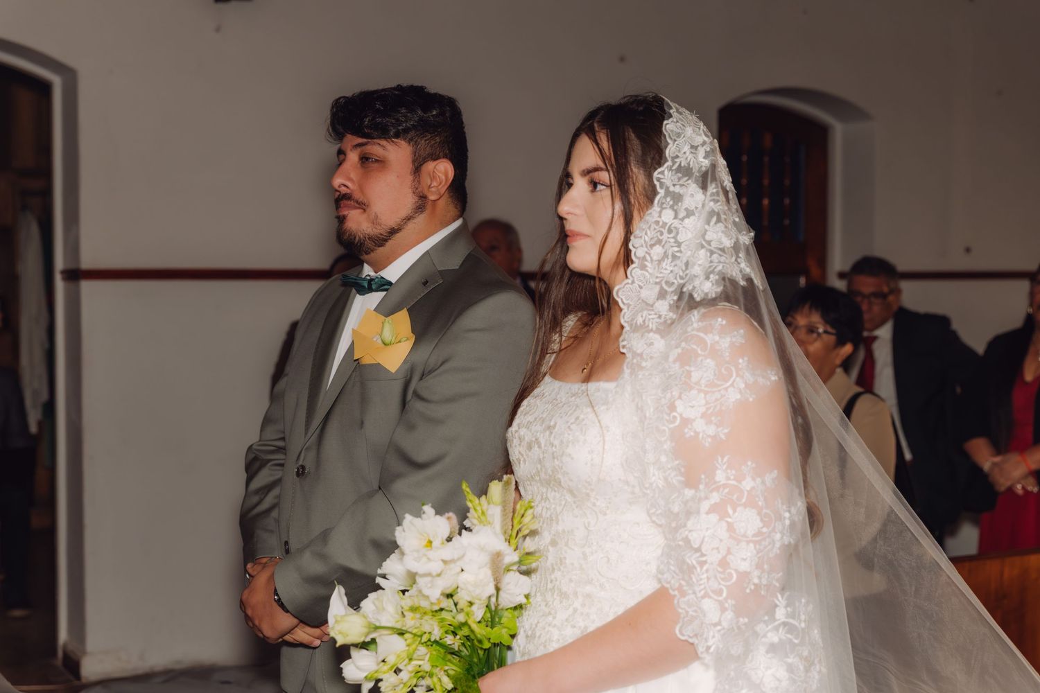 Bride in white lace veil and groom in gray suit stand together at church wedding ceremony.