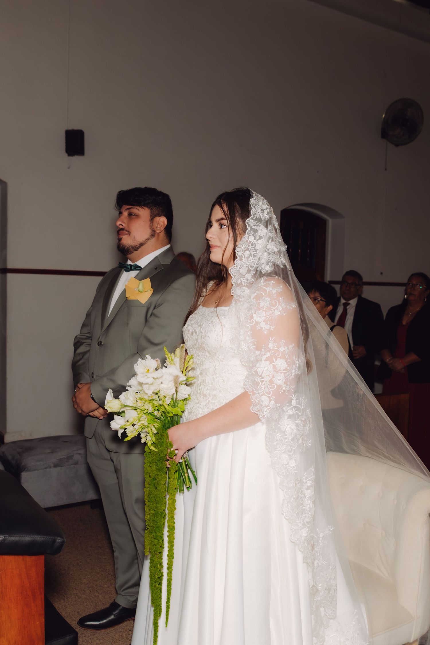 Bride in white lace gown and veil holds bouquet beside groom in gray suit during church wedding ceremony.