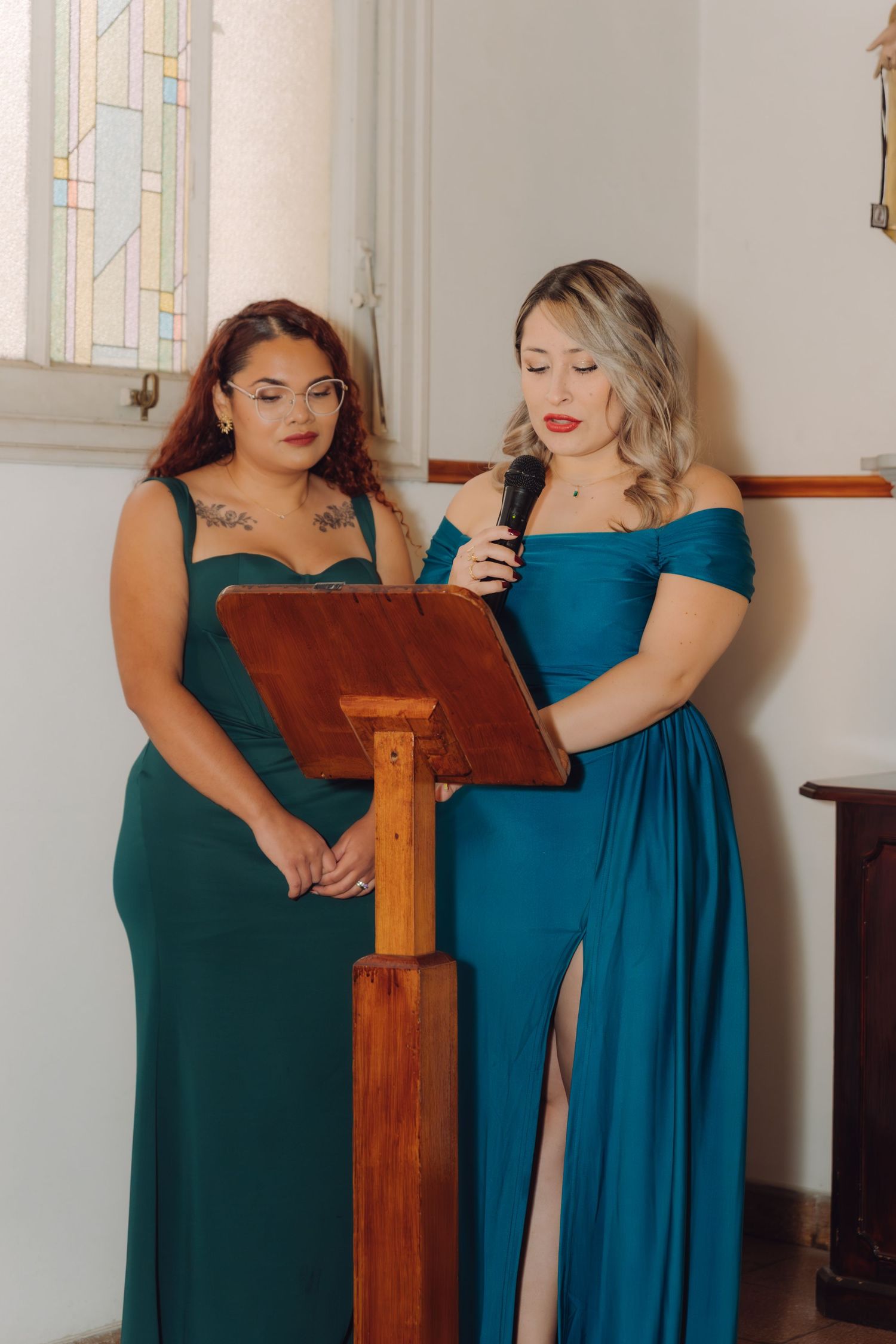 Two women in teal evening gowns read from a lectern with microphone during a wedding ceremony.
